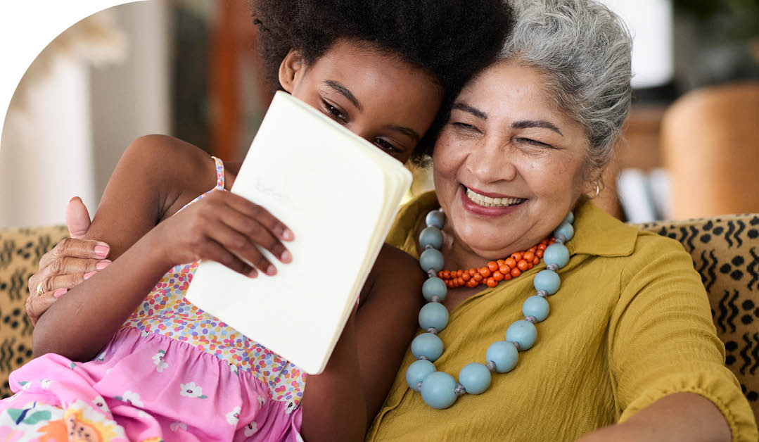 Girl reading book while sitting with grandmother on chair at home